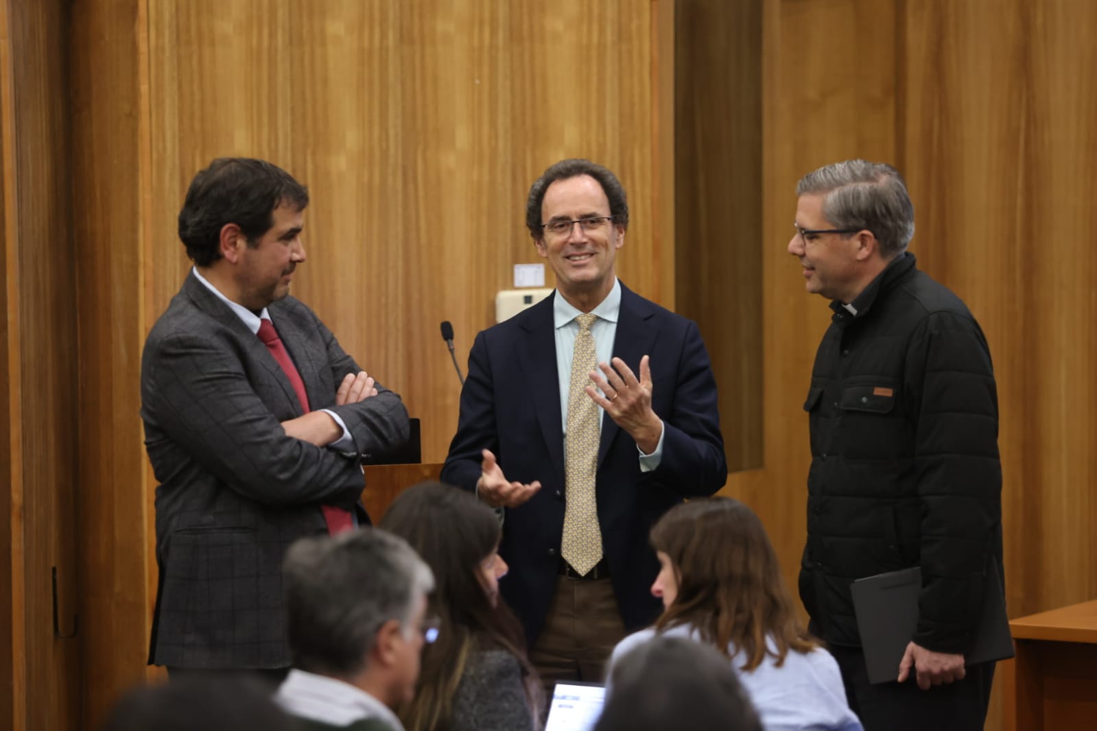 La imagen muestra a tres hombres conversando de pie en un entorno interior, probablemente un auditorio o sala de conferencias, con paneles de madera al fondo.